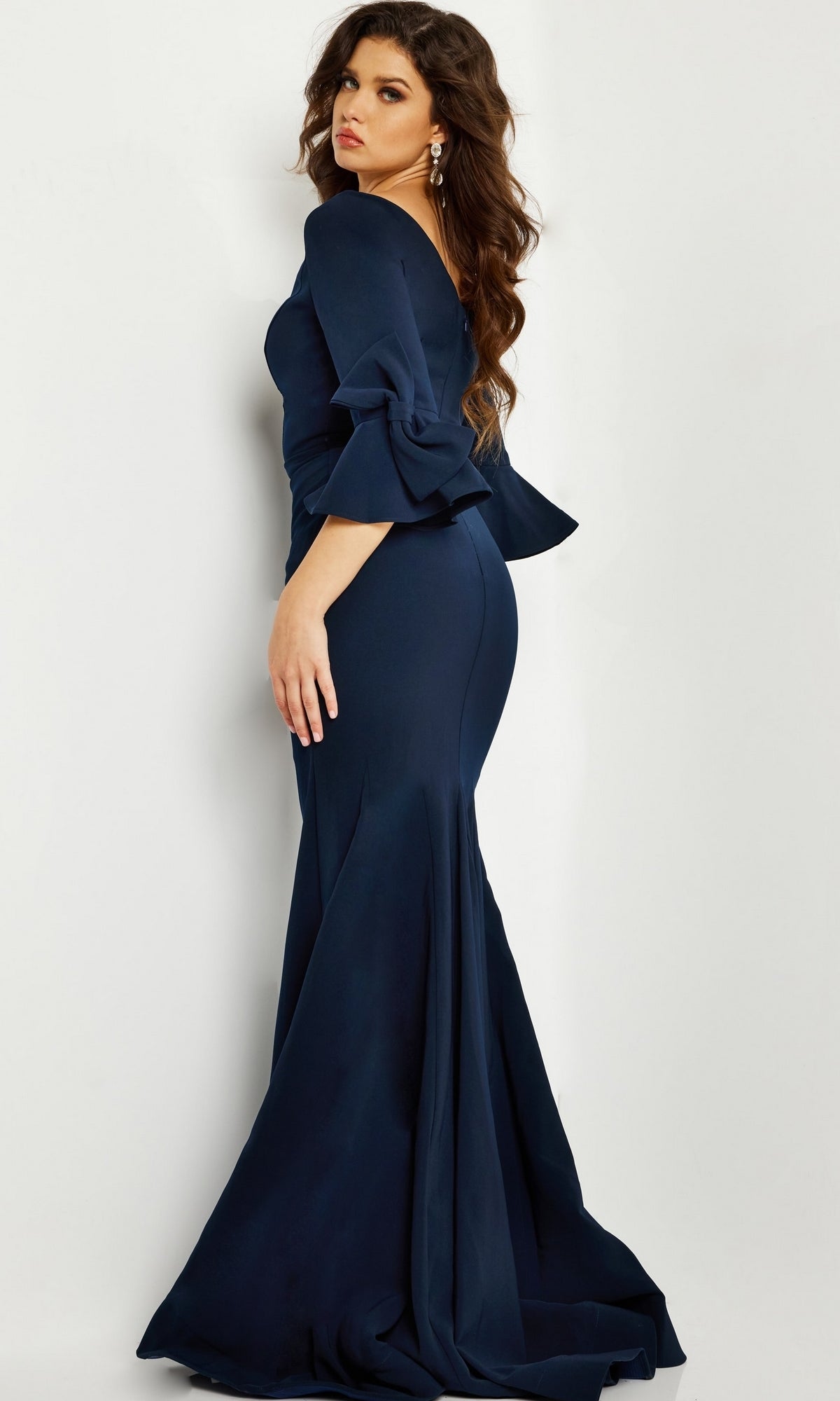 A woman models the Long Formal Jovani Dress 09776—a fitted, floor-length navy gown with flared sleeves—standing sideways and looking over her shoulder against a plain light background.